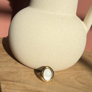 Bague dorée de style vintage en acier inoxydable sertie d'un grand cabochon ovale en nacre blanche irisée, posée sur une table en bois clair près d'un vase texture grès.