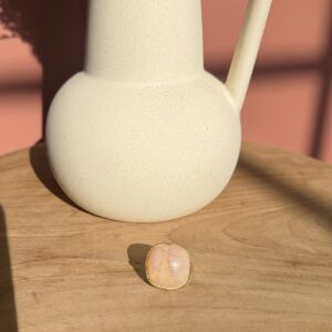 Bague dorée en acier inoxydable avec une grosse pierre quartz rose ovale et lisse, posée sur une table en bois clair devant un vase blanc.