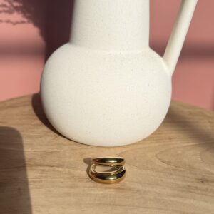 Bague double anneau minimaliste en métal doré lisse, design bombée et moderne, posée sur une table en bois clair.