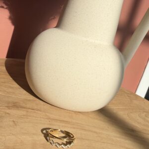 Bague ajustable dorée à double rang, composée d'un anneau lisse et d'un anneau en zigzag serti de strass brillants, sur un support en bois.
