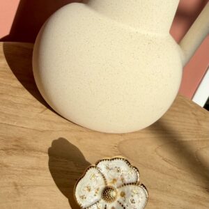 Bague en forme de fleur ciselée blanche et dorée avec des inclusions de feuille d'or, posée sur une table en bois clair près d'un pichet texturé