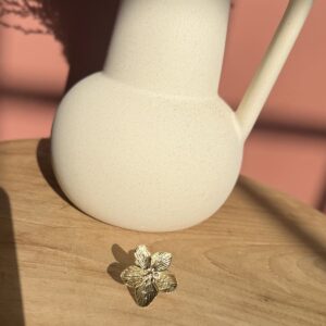 Bague dorée en forme de fleur ciselée à 5 pétales, posée sur une table en bois clair près d'un pichet texturé.