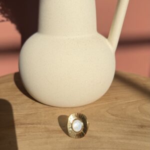 Bague dorée de style vintage avec un motif soleil gravé et une nacre blanche au centre, posée sur une table en bois clair près d'un pichet texturé.