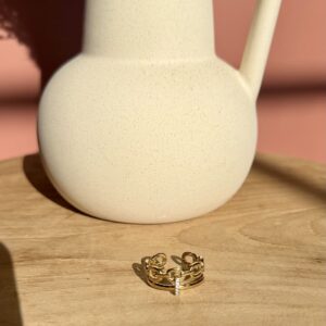 Bague double anneaux dorée style maillon de chaîne avec pendentif barre serti de cristaux sur un socle en bois."