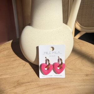 Boucles d'oreilles créoles dorées Milémila en acier inoxydable avec une breloque en résine rose fuchsia vif en forme de cœur, présentées sur leur carte de marque.