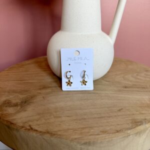 "Paire de boucles d'oreilles dorées en acier inoxydable avec pendentifs en forme de fleurs, présentées sur une carte de la marque Milë Mila posée sur un tabouret en bois."