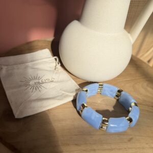 Bracelet Milémila composé de perles tubulaires en résine bleu ciel effet marbré, séparées par des anneaux dorés en acier inoxydable, présenté avec son pochon sur un support en bois.