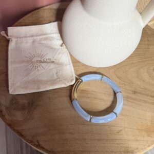 Bracelet Milémila en perles acryliques tube bleu ciel marbré, séparateurs et un grand segment dorés en acier inoxydable, posé avec son pochon sur une table en bois.