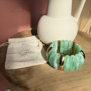 Bracelet élastique Milémila composé de segments en résine vert d'eau et d'inserts dorés, présenté avec son pochon en tissu sur un plateau en bois clair.