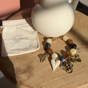Bracelet Milé mila en perles de résine marbrées (ambre, noir et crème) avec breloques dorées scarabée égyptien, abeille et cœur, à côté de son pochon en lin.