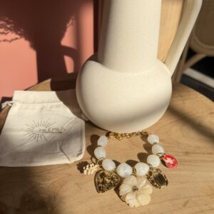 Bracelet de perles blanches Milémila avec breloques dorées (cœur, fleur, scarabée, fraise) et pochon en lin, posés sur une table en bois clair devant un vase texturé.
