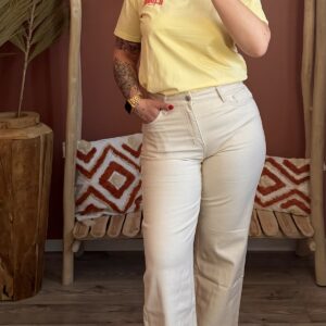 Femme portant un t-shirt jaune pâle avec inscription rouge, associé à un pantalon large écru ou beige clair et des sandales beiges, dans un décor intérieur bohème.