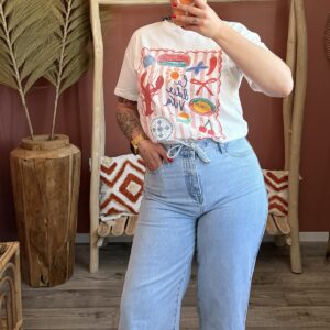 emme portant un t-shirt blanc à imprimé graphique "La dolce vita" coloré, associé à un jean large bleu clair à taille élastiquée et des sandales beiges.